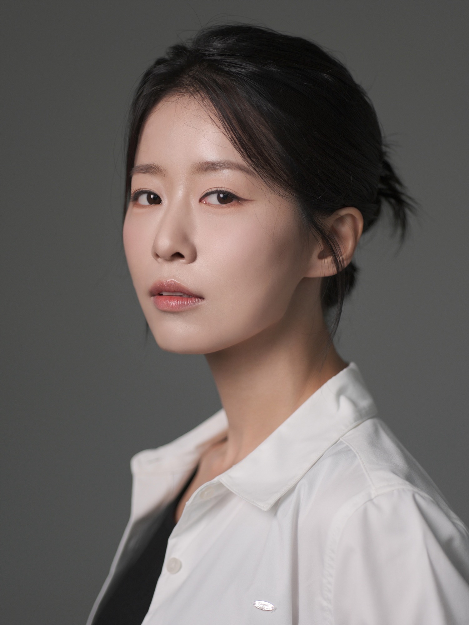 회색 배경에서 흰 셔츠를 입고 묶은 머리로 정면을 보는 여성 배우 프로필
