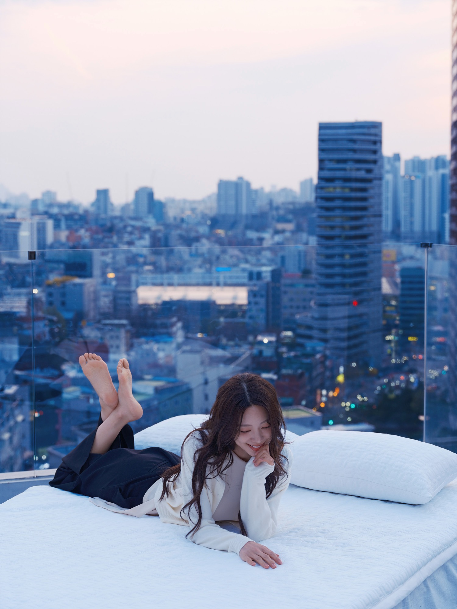 해 질 무렵 도심 빌딩 옥상에 침구를 펼치고 여성 모델이 앉아 있는 브랜드 캠페인 사진