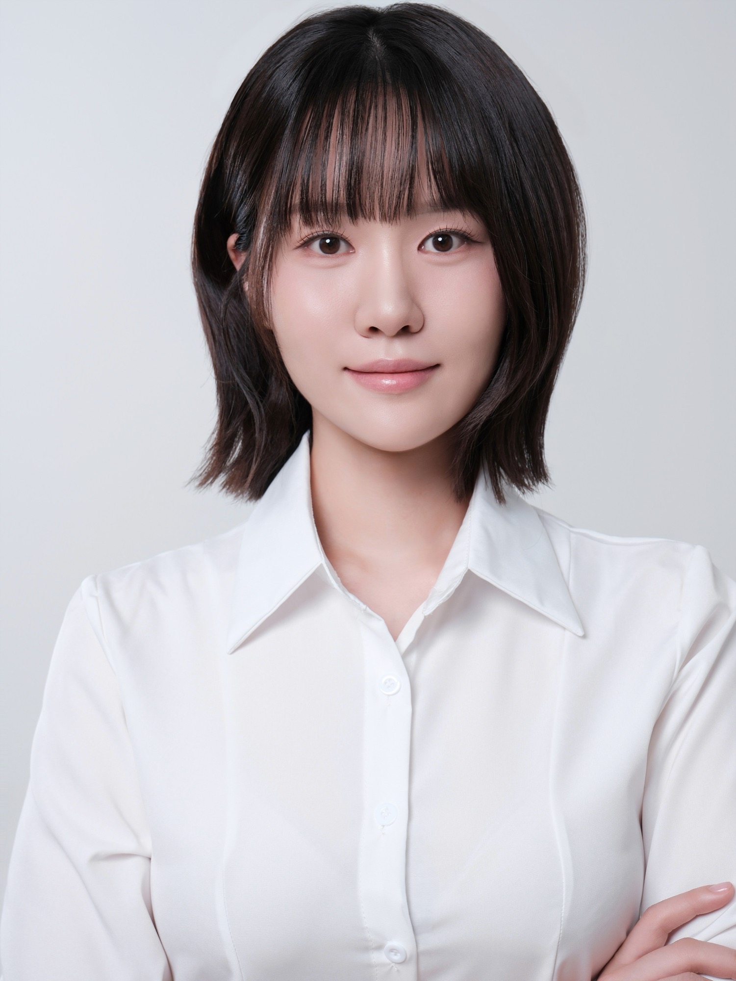 회색 배경에서 흰 셔츠를 입고 팔짱을 낀 단발 여성이 입꼬리만 살짝 올린 정면 컷