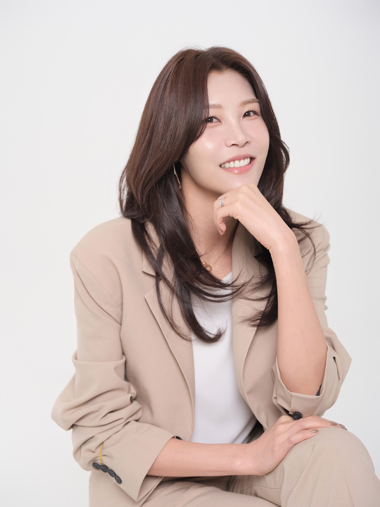 밝은 배경에서 베이지 정장을 입고 앉아 턱에 손을 대고 미소 짓는 여성 프로필