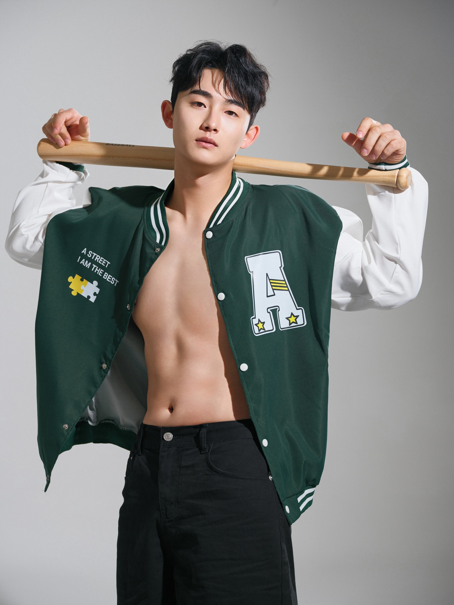 회색 배경에서 스타디움 재킷을 입고 야구 배트를 어깨에 걸친 남성