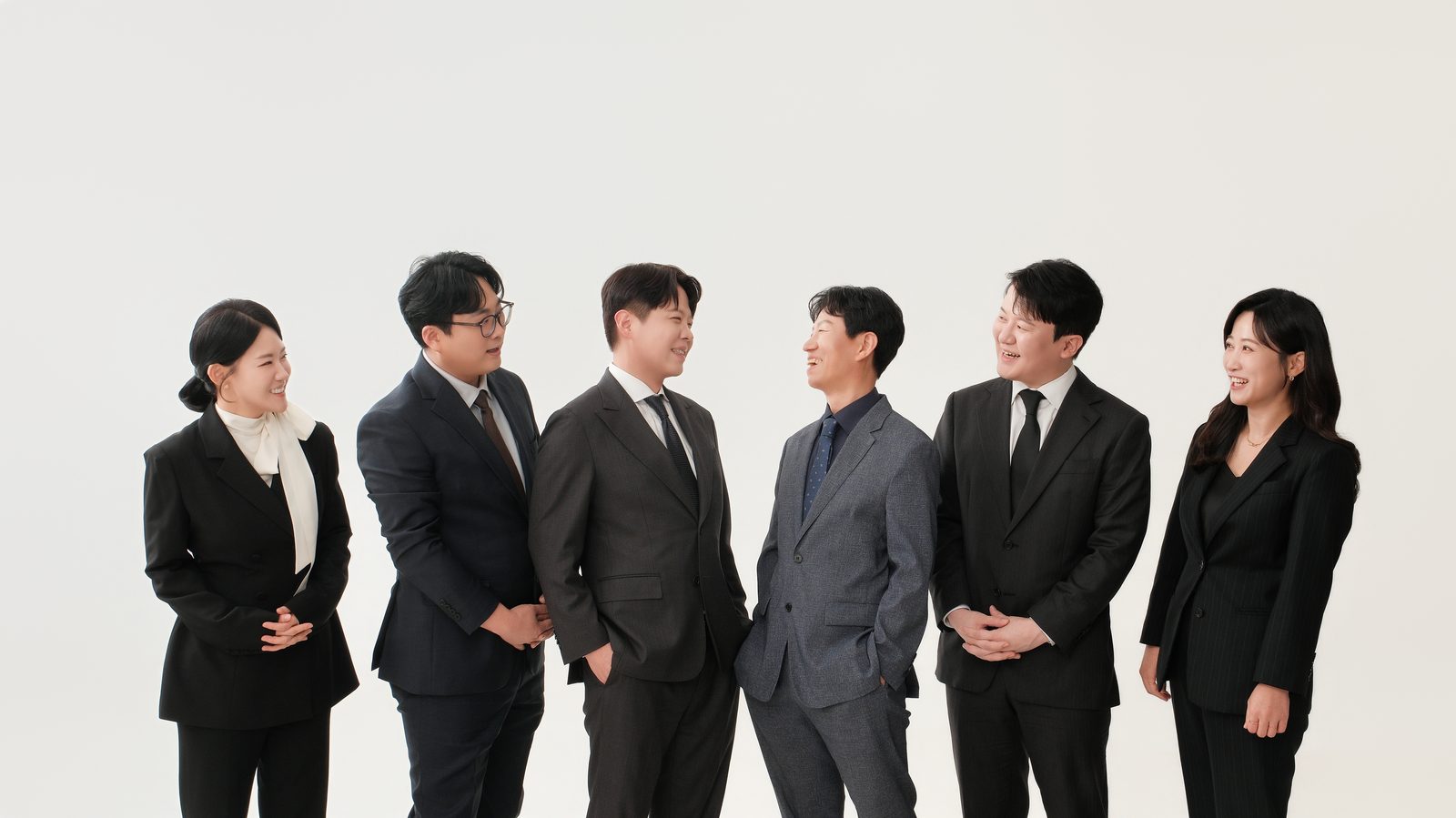 정장을 입은 직원 여섯 명이 흰 배경에서 서로를 바라보며 서 있는 단체 프로필 사진